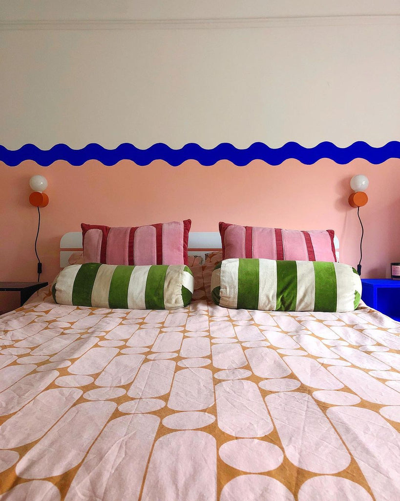 Bedroom with striped bedding and pillow covers, photographed in front of a three-coloured wall: neutral, Electric Blue and peach