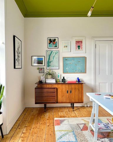 neutral coloured wall with olive green coloured ceiling, decorated with a gallery wall with a variety of art prints, a mid-century wooden cabinet and a natural wood floor