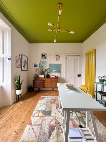 photograph of a home office with neutral walls and a big window on the left with natural light coming in, an olive green ceiling, bright yellow radiator on the right, dark wooden floor, gallery wall and a plain white desk placed on the right side