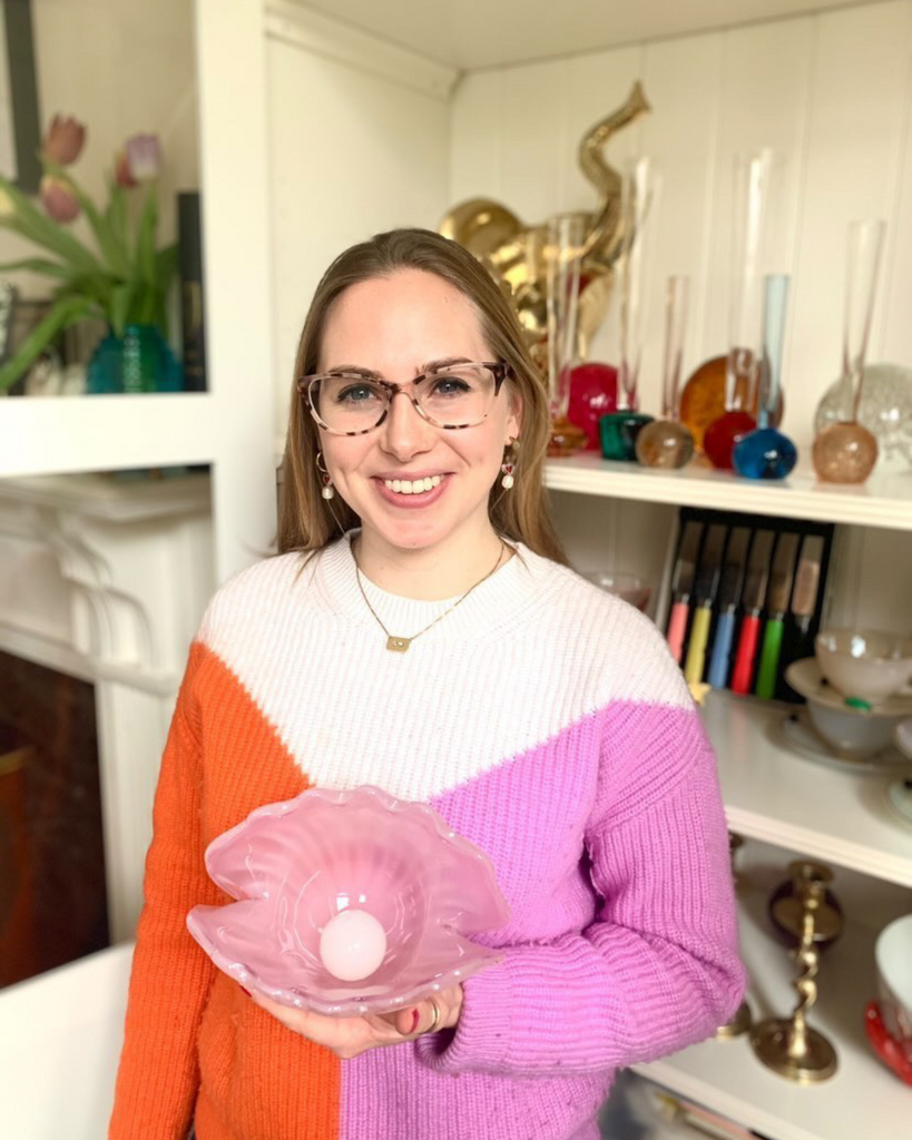 Blonde woman with glasses and a knitted jumper in white, pink and orange, photograph in front of a white wall with white shelves, and holding a shellfish vintage glass item