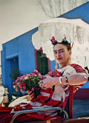 A photograph of artist Frida Kahlo dressed in a red dress, holding red flowers and sitting on a chair while keeping a shade with a white parasol, while in the background is her blue house painted in a bright blue colour with red door and window frames