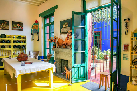 Photo of a traditional rustic kitchen in a bright yellow colour, including a big wooden dining table and many plates and ornaments hanging from the walls, leading to teal garden doors with small windows, showing the view to a big garden with greenery and bright blue walls