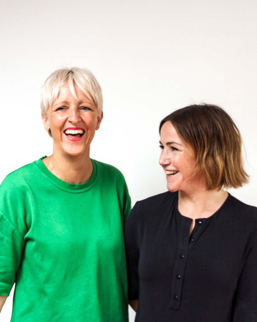 a tall blonde woman with short hair and green t-shirt photographed next to a shorter woman with shoulder-length hair and black t-shirt in front of a white wall