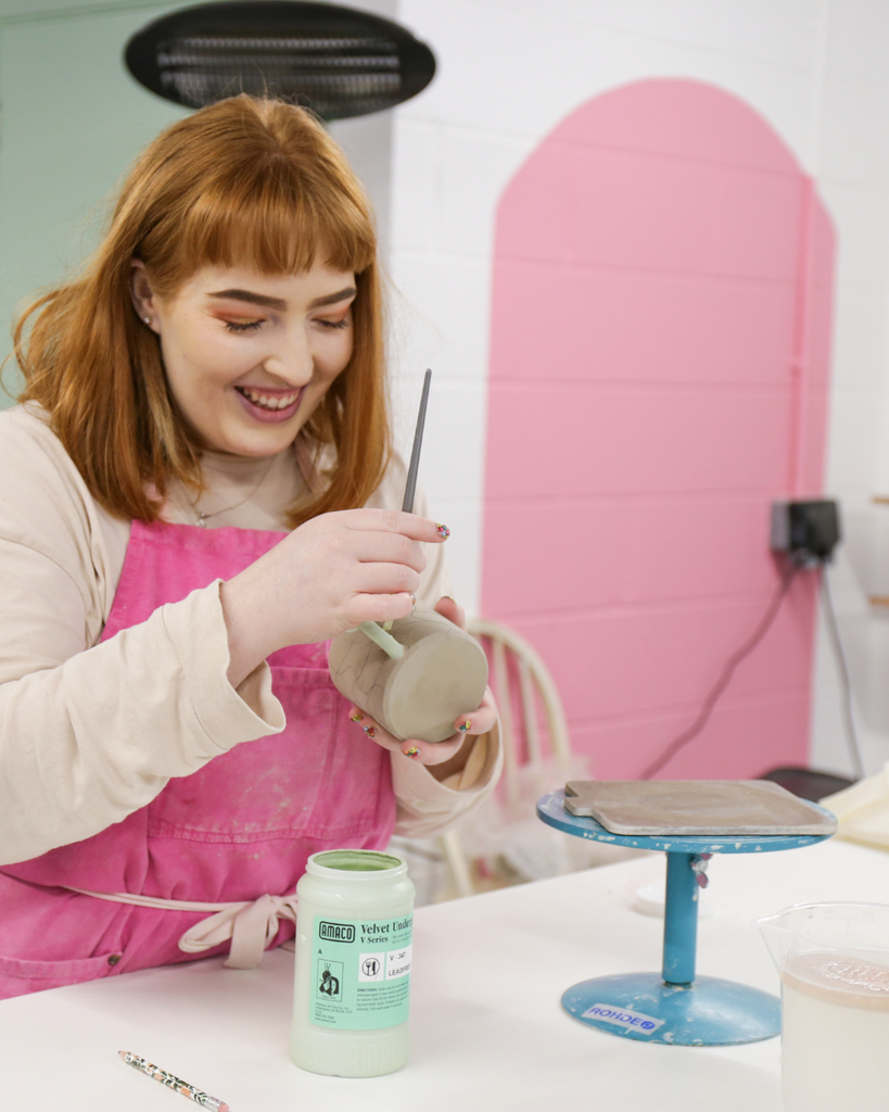 white woman with ginger hair and hot pink apron working on a ceramic piece, photographed in front of a white wall with a pink paint zoning area