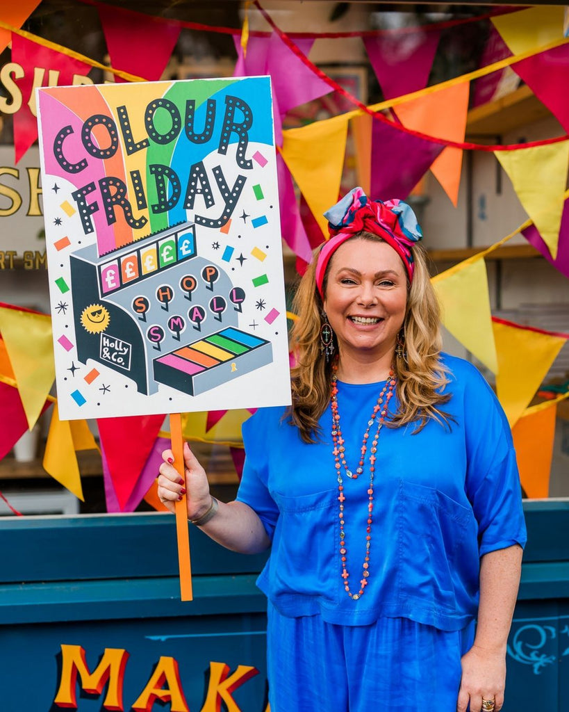 A dark blonde woman in a blue two-piece outfit holding a colourful sign that says 'Colour Friday'