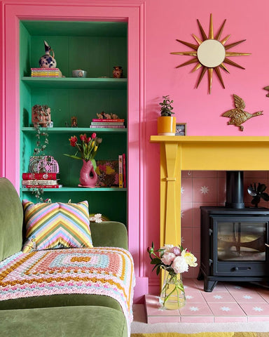 photo of the main wall of a living room with the wall painted in bubblegum pink colour and the shelving in bright green, next to that a yellow fireplace and a forest green sofa to the left, decorated with knitted cushions and throws