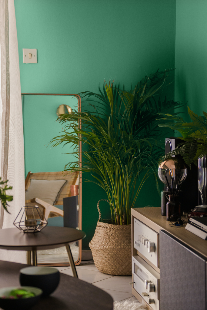 A green-teal coloured bedroom with a large plant placed in the corner, a large standing mirror next to it and a neutral sideboard on the right side, decorated with minimal home furnishings