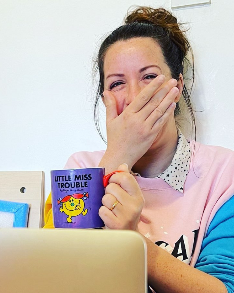 Brunette woman laughing in front of a computer while holding a purple coffee mug that says 'Little Miss Trouble'