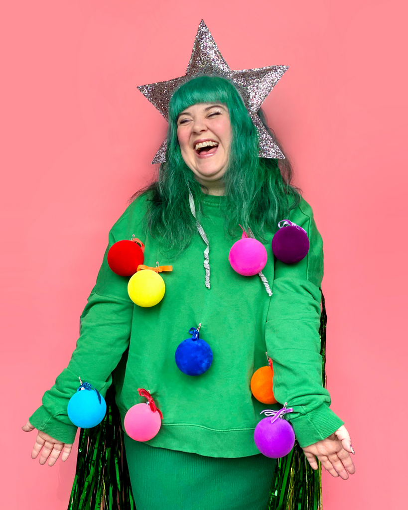 woman with green Christmas tree costume with colourful pom poms and star hat, photographed in front of bubblegum pink coloured wall