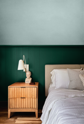 photo of a pale coloured bed placed on the right side of the frame, next to a vintage wooden mid-century style bedside table and a small table lamp placed on top of it next to a little stone sculpture, all photographed in front of a split coloured wall where the top part is a pale grey green colour and the bottom part is a dark green colour