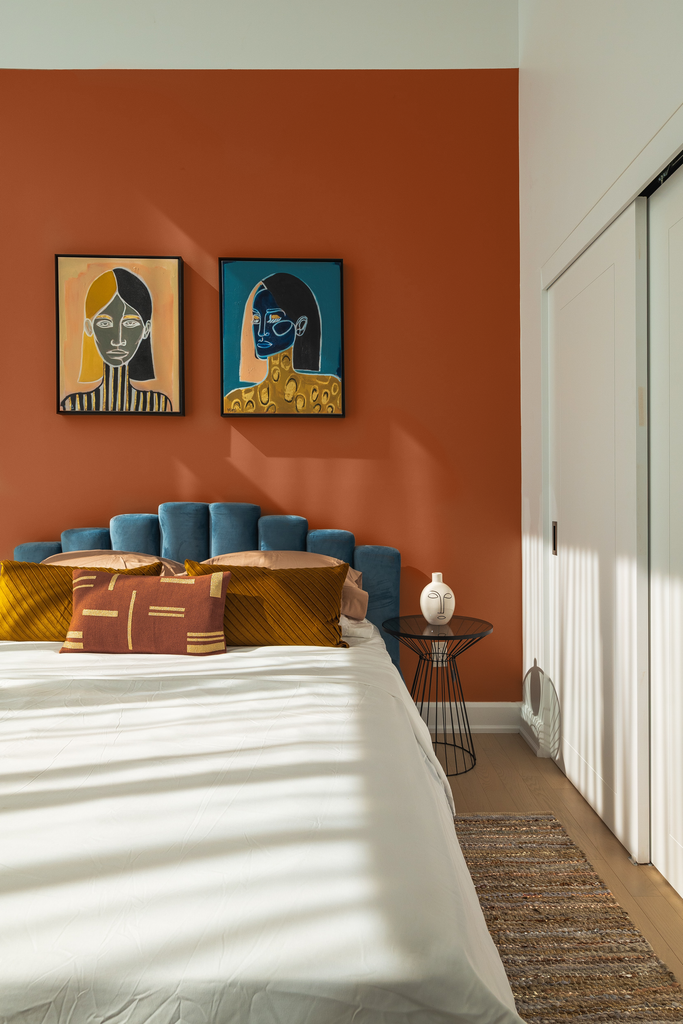 Double bed with blue velvet bed frame photographed in front of rusty dark orange paint colour with two African art-inspired artworks hanging on the wall above the bed