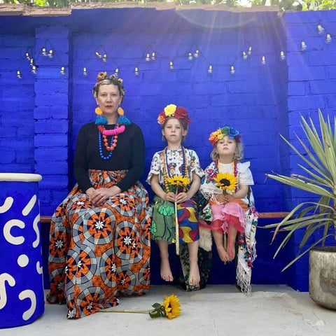 A photograph of a blonde woman and two blonde girls, sat on a bench with colourful dresses and flower crowns, surrounded by tall plants and posing in front of a ultramarine blue brick wall