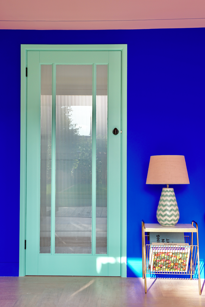 Area of a room with a ceiling painted in peachy pink colour, wooden door frame in mint green and wall in electric cobalt blue, with a magazine rack and a table lamp placed on the right side of the door