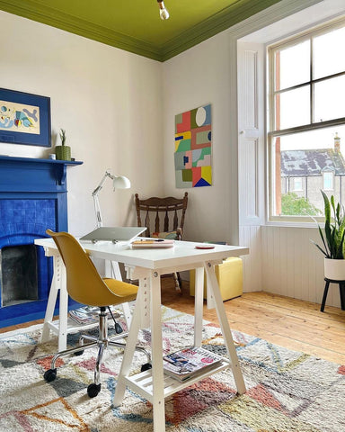 Home office with blue painted fireplace, olive green painted ceiling and neutral walls; white wooden desk and yellow chair, light coloured rug and colourful artworks hanging on the walls