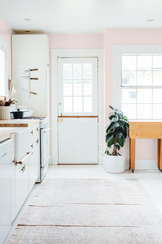 photo of a spacious neutral kitchen with a lot of natural sunlight, on the left there are white kitchen cupboards and a fridge, then a neutral vintage rug on the floor and a peach coloured wall next to the back yard door and window frame