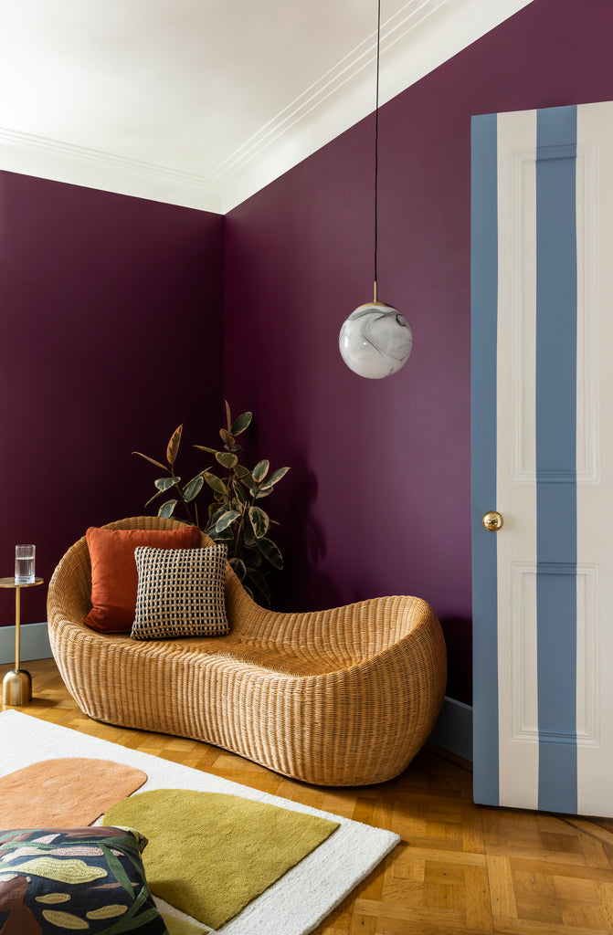 A corner photo of a rustic living room with a white and blue stripes painted on the wall, white ceilings, dark purple Viva Magenta painted walls, rustic rattan sofa, olive tree and home furnishings in warm autumn colours