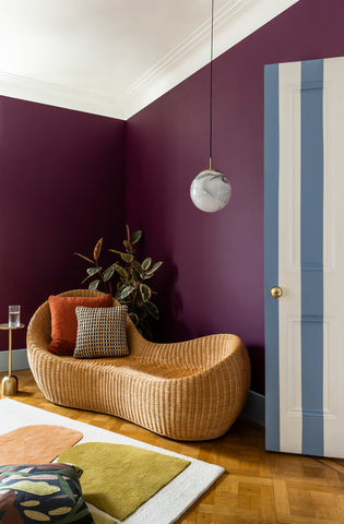 photo of a living room with a white ceiling and dark purple pink walls, with blue and white stripes on the door on the right, dark wooden floor, earthy green and orange coloured warm rug and rattan daybed, decorated with orange cushions and plants