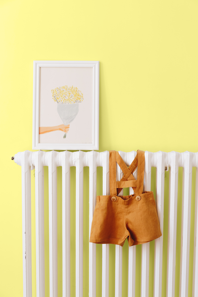 Nursery room with walls painted in bright pastel yellow paint colour, with big radiator, colourful watercolour art print and a children's one-piece outfit hanging on it