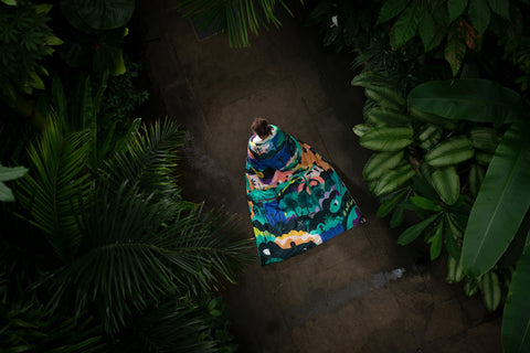 an image of a kid with a colourful blanket hanging from its back, walking on a path between two green bushes in a botanical nature setting 