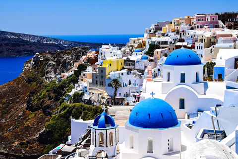 landscape photo of Santorini with a bright blue sea, a dark green mossy hill on the left and a layered line of white houses and church buildings with ultramarine blue circle domes on the right