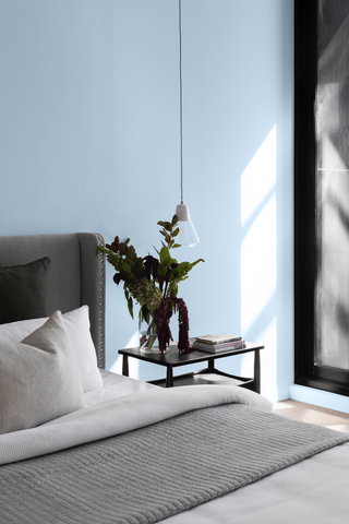 photo of a grey bed photographed on the bottom left corner of a light blue coloured wall, with white and bed bedding, a wooden bedside table and a hanging plant placed on top of it, with natural light coming in from a window on the right
