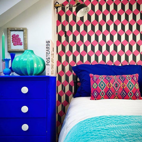 bedroom photo of a bed placed on the right side with one ultramarine blue and one middle-eastern inspired pattern pillows, white sheet and aqua coloured throw, photographed in front of a geometric-shaped wallpaper in red and white and a bedside table painted in a ultramarine blue colour and decorated with a large circle teal vase on top