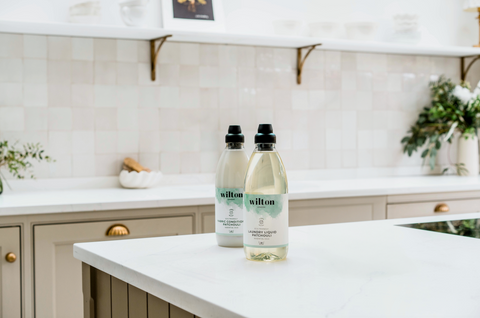 photo of a spacious contemporary kitchen with light beige square tiles, light brown coloured cupboards and a kitchen island with a white countertop, with two plastic bottles filled with liquid placed on top of it