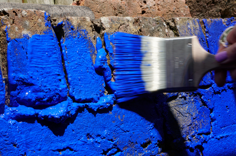 A brick wall with a hand holding a wide paint brush on the right side while painting the bricks in ultramarine blue colour in one motion