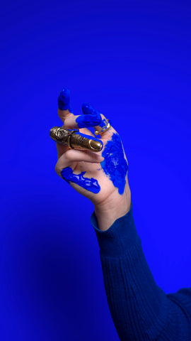 photo of an extended hand with a dark royal blue long sleeve, photographed in front of a Yves Klein blue background, with ultramarine blue paint splashed all over it and a golden all-finger ring on her finger