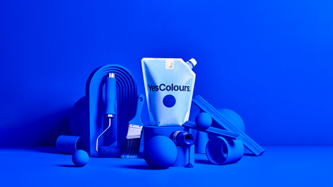 A photograph of multiple home decor and DIY objects - paint roller, paint brush, as well as small objects in various shapes, all painted in ultramarine blue colour and arranged around a white pouch with logo 'YesColours', all photographed in front of an ultramarine blue background