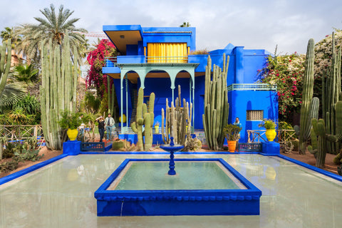 Majorelle Garden's fountain in light green colour and painted frame in ultramarine blue colour, photographed in front of a ultramarine blue and mint green palace surrounded by tall cacti and palm trees