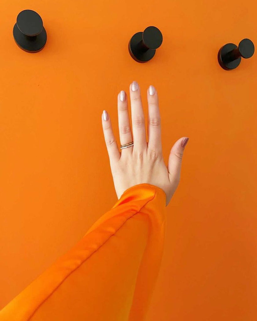 A pale white hand with an orange dress sleeve photographed posing in front of a bright orange-painted bathroom wall with black hangers above it