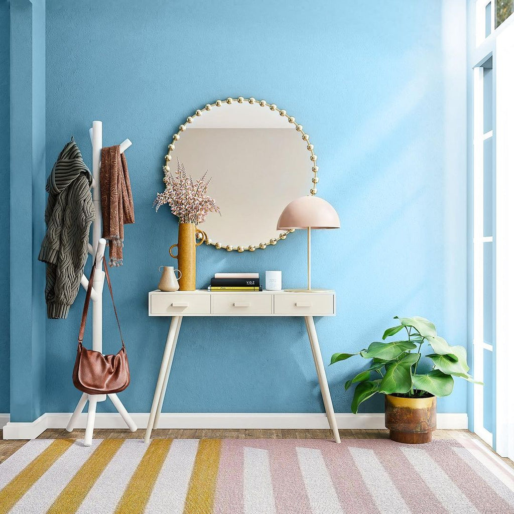 A spacious, airy hallway with a striped white, mustard yellow and pale pink rug, with a little sideboard with flowers on top, hanging mirror above it, clothing rack on the left and small potted plant on the right, all photographed in front of a pastel blue wall