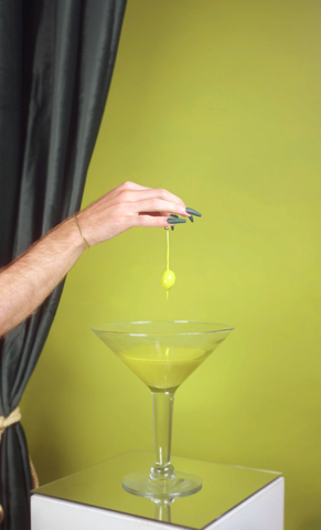 photo of olive green wall with a dark velvet green curtain showing on the left, with a pale skin hand coming out of the corner with dark green long nails, holding an olive that's dripping green paint onto a cocktail glass