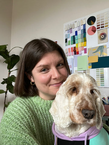 photo of a woman with long bob haircut and green jumper, photographed in front of a neutral wall with a tall plant and paper colour cuttings and holding a sand coloured spaniel dog dressed in a colourful lilac, black and aqua jumper