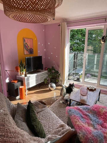 photo of a living room with pink walls and orange coloured mural, with a disco ball on the wooden floor reflecting the natural light on the walls, grey sofa and colourful pink and blue throw