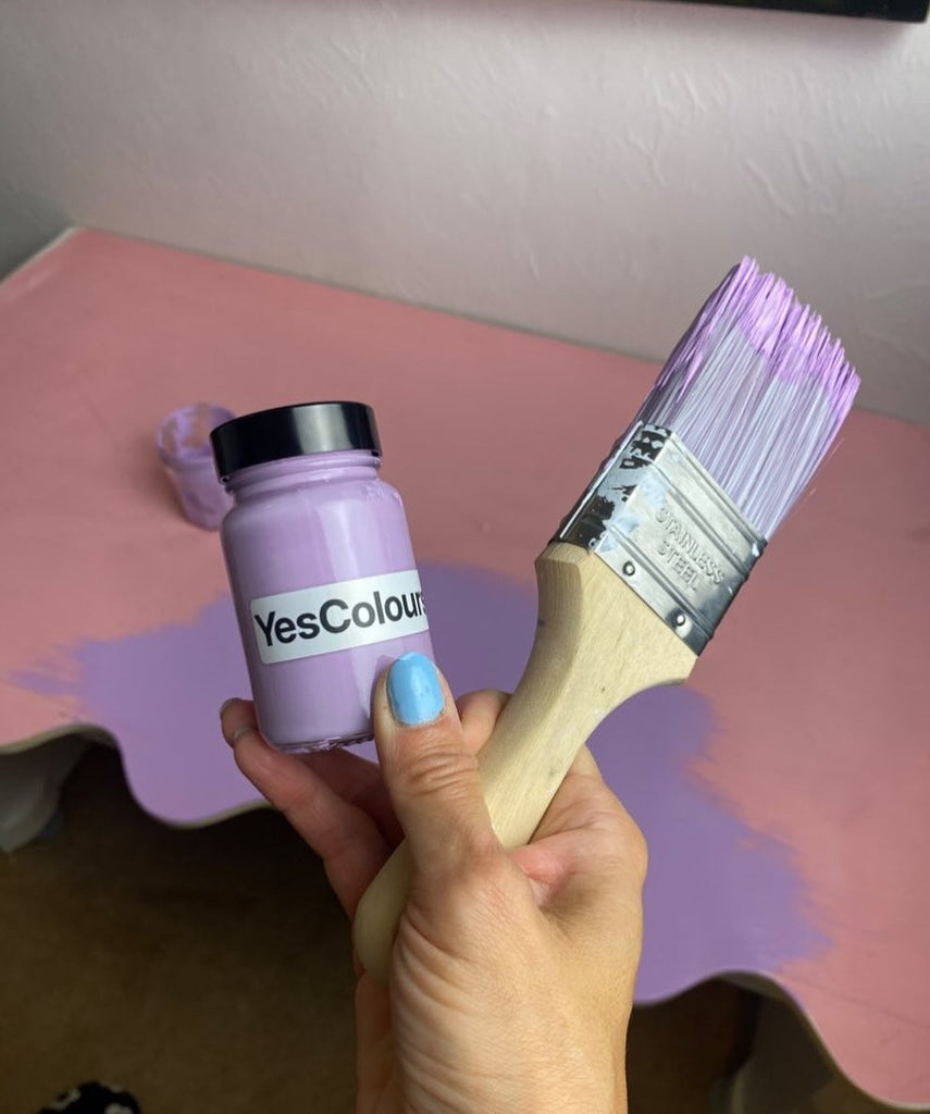 A female hand with blue nails holding a YesColours Joyful Lilac paint sample in a glass jar with a paint brush dipped in lilac paint