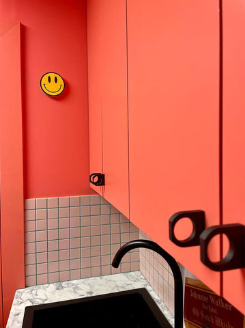 close up of a kitchen cupboard area with three visible kitchen cupboard painted in a coral orange colour, with added matt black handles, supported by a small area of square tiles and a matt black sink