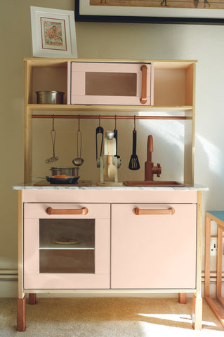 a children's play kitchen toy structure with peachy coloured cabinets and mini stove, with toy kitchen utensils hanging from the top shelf and photographed in front of a dark neutral wall