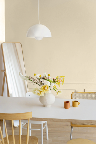 image of a dining room corner with cream-coloured walls, a big standing mirror on the left, hanging white pendant light, a long rectangular white dining table, two chairs on its sides and a bouquet with yellow spring flowers placed in a vase