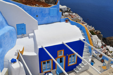 Photo of an island architecture consisting in a bright ultramarine blue painted house with orange window frames and white rooftop, surrounded by a bright blue stone wall, overlooking a blue sea to the right