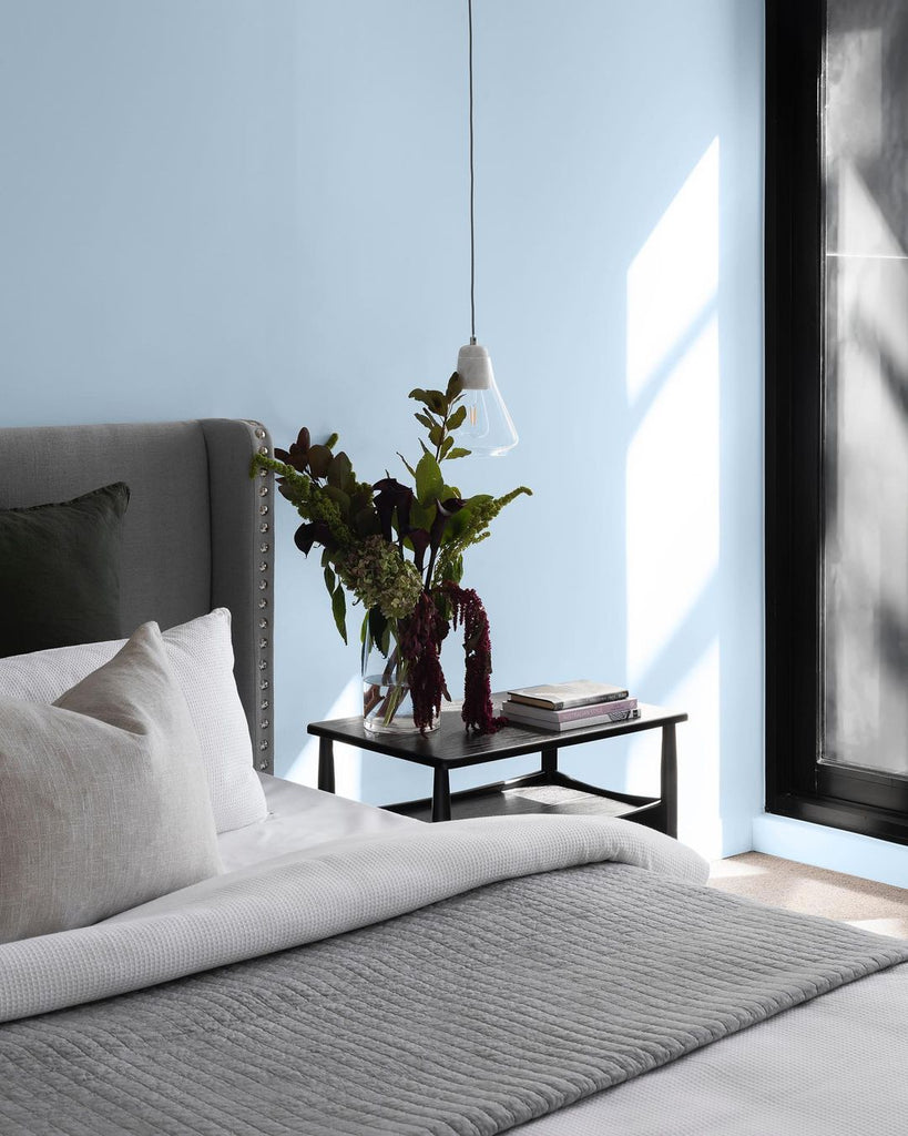 Bedroom corner with a hanging lightbulb and grey bed headboard, photographed in front of a pale blue wall