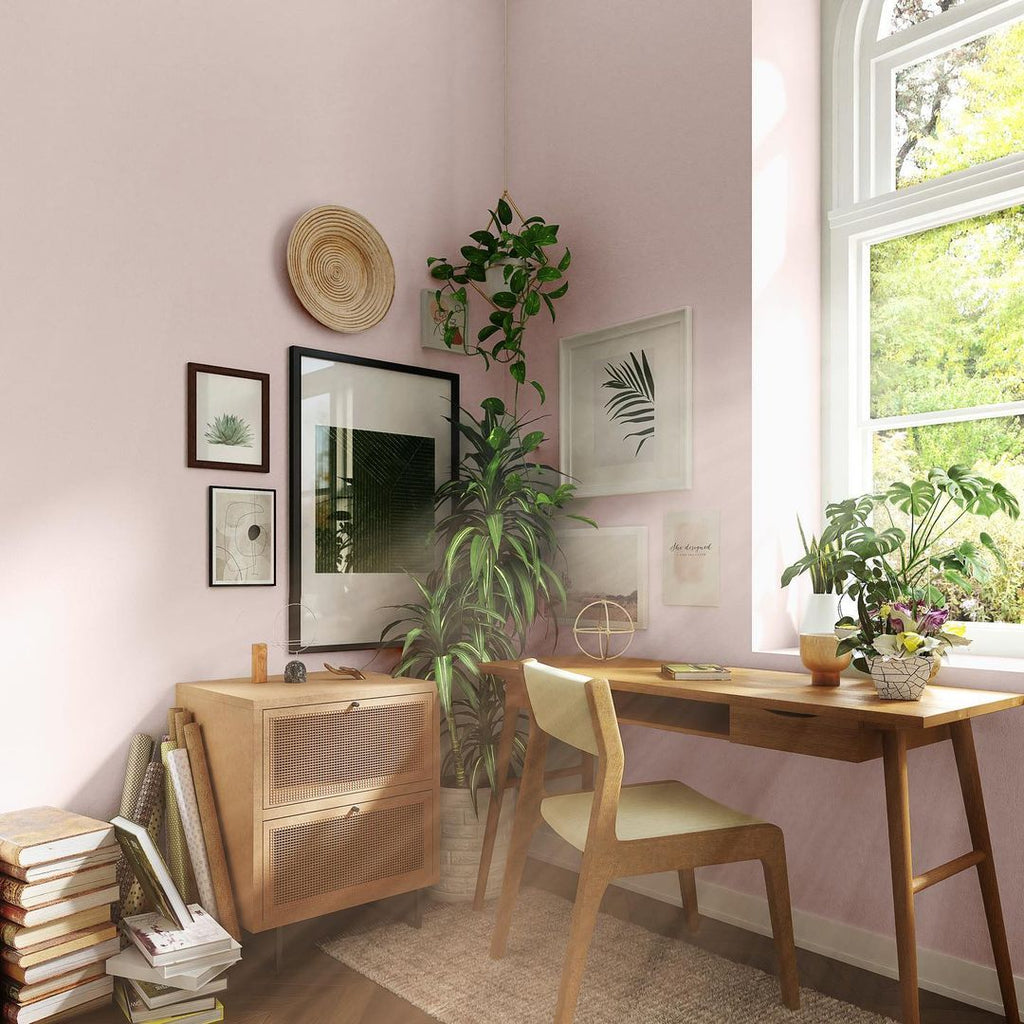 Home office area painted in light peach paint colour, with wooden mid-century design desk and chair, sideboard, hanging plans and a stack of books placed on the floor
