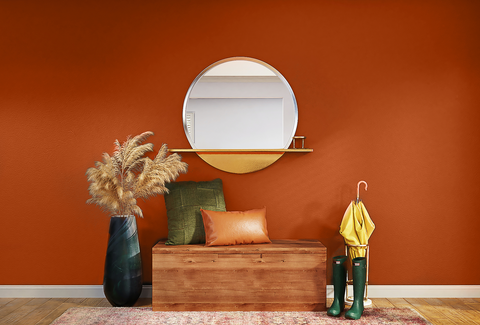 photo of a large rusty orange terracotta wall, with a wooden bench placed on top of it, decorated with green and orange cushions, pampas grass on the left and rain boots and umbrella to the right, with a round mirror above it