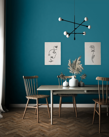 Dining room scene with the wall painted in a dark teal colour, with wooden dining table and two chairs arranged around it, two vases with pampas grass placed on top of the table, geometric chandelier and two black and white illustration artworks on the wall