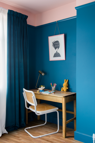 photo of a children's bedroom cover with a vintage wooden desk, white and rattan chair as well as a plush bunny toy, crayons and a table lamp placed on top of it, surrounded by a dark teal colour on the walls and a peachy pink colour on the ceiling
