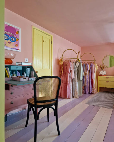 Pastel coloured bedroom with peachy pink walls, bright yellow door and striped wooden floor in lilac and white, decorated with yellow bedside table, rattan chair and colourful dresses hanging from a clothing rack