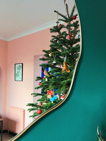 photo of a green Christmas tree next to a peach coloured wall, photographed in the reflection of an asymmetrical mirror with a golden frame placed on a dark green painted wall