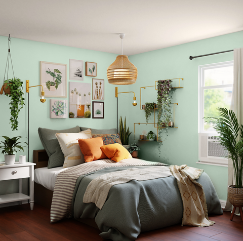 light green coloured bedroom with a neutral ceiling, with seven botanical artworks placed on the main wall, surrounded by leafy plants and a grey and white queen sized bed with grey and bright orange cushions
