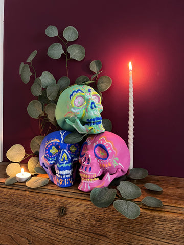 image of three artificial skulls in blue, green and pink and with wavy lines and dots decorated on them, stacked on top of each other next to a tall candle, small tea candle, leafy plant, all photographed in front of a magenta coloured purple pink wall